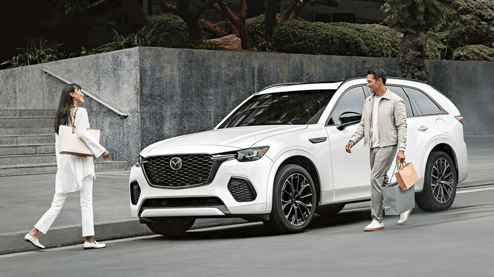 vue latérale avant d'une Mazda CX-70 garé sur le bord d'une rue près de son conducteur et une passante